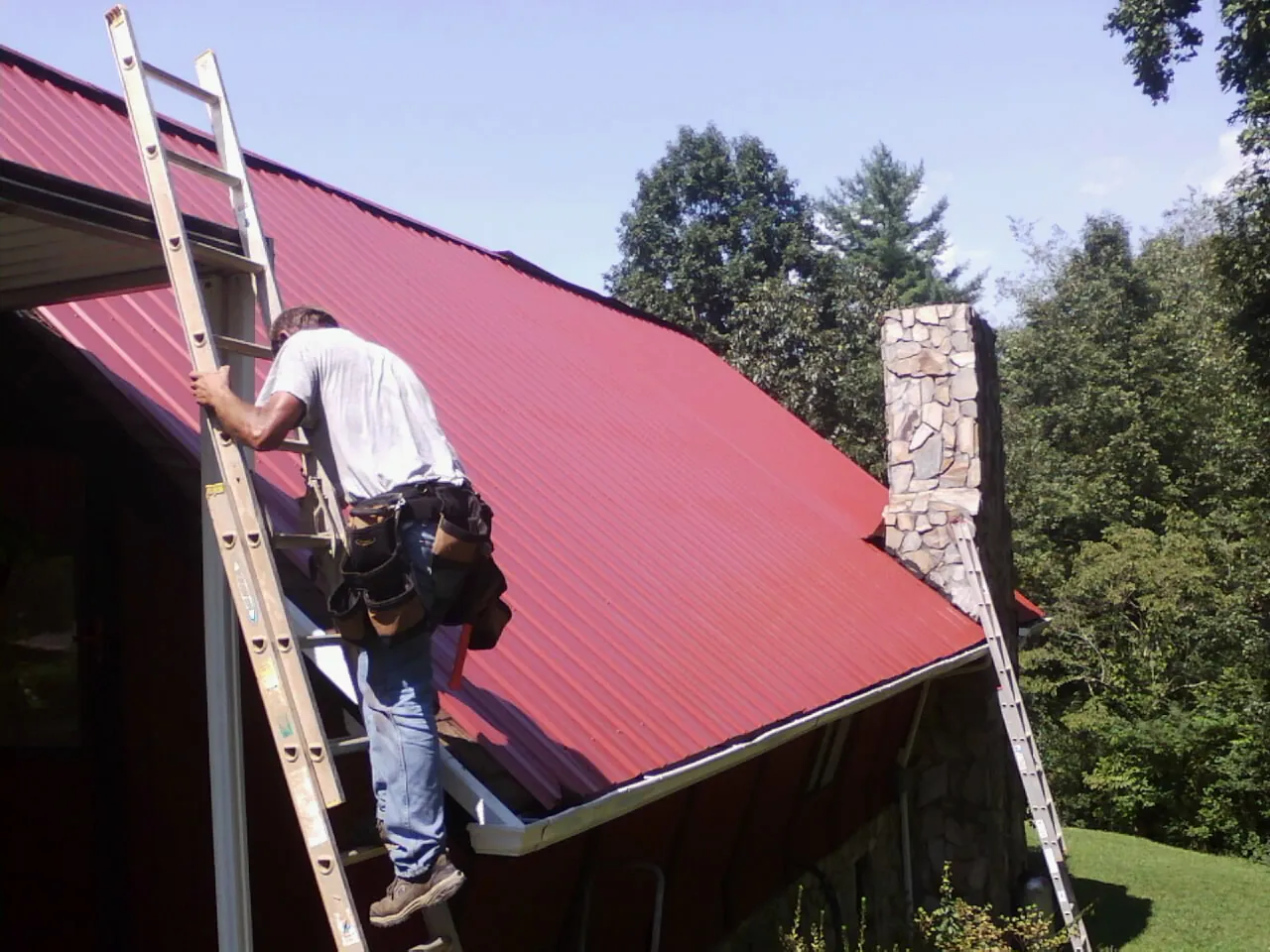 Roofer on a ladder beside a red metal roof and stone chimney — Soffit & Fascia Repair by Roofing Brothers in Mount Pocono